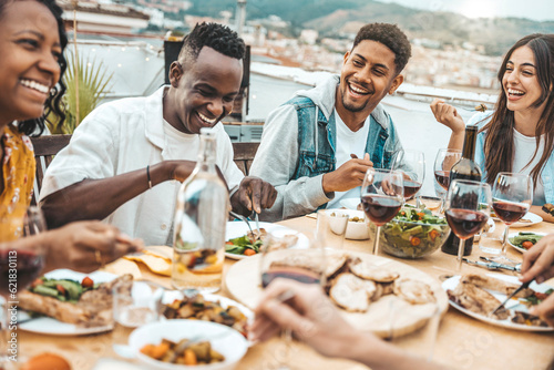 Multiethnic friends having fun at rooftop bbq dinner party - Group of young people diner together sitting at restaurant dining table - Cheerful multiracial teens eating food and drinking wine outside