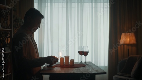 The man is getting ready for a date. The dark silhouette of a man lights candles with matches. Candles and glasses with red wine on the table.