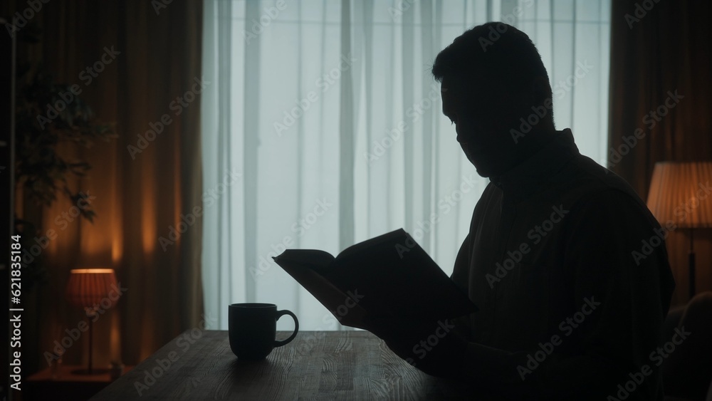 A man reads a book sitting at a table with a cup of coffee, tea in the room on the background of the window. Dark silhouette of a man with an open book in his hand close up.