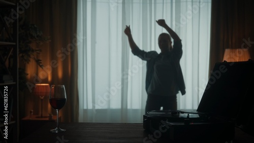 Vintage record player with a vinyl record and a glass of red wine on the table close up. Carefree woman dances cheerfully alone near the window. Dancing woman silhouette in the living room.