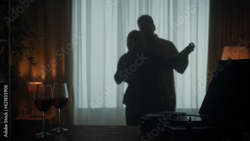 Vintage record player with a vinyl record and glasses with red wine on the table close up. A young couple is dancing a slow dance while enjoying a pleasant evening together. Silhouette.