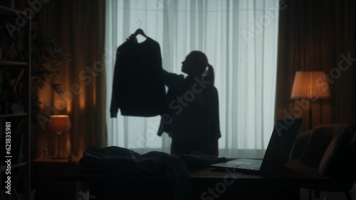 A young woman chooses what to wear. A woman holds a blouse on a hanger in her hand and examines it carefully in order to make the right decision. Dark silhouette of a woman in the living room.