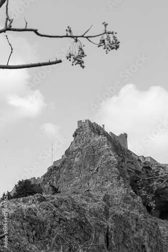 View from Saint Paul's Bay of the Acropolis of Lindos fortress on top of a mountain with tourist person standing at the top in black and white