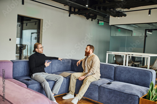businessman in black turtleneck and eyeglasses pointing with finger while talking to bearded colleague on soft couch in contemporary office, partnership and success concept