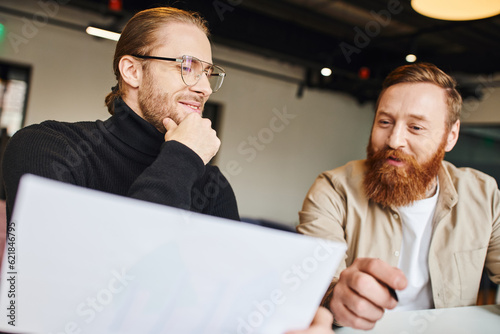 pleased businessman in black turtleneck and eyeglasses touching chin while looking at document near happy bearded colleague while working on new startup in coworking office