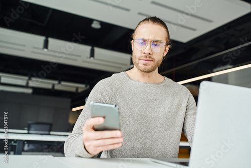 entrepreneur in eyeglasses and casual clothes, with serious face expression, looking at mobile phone while working on startup project near laptop on blurred foreground in modern office space