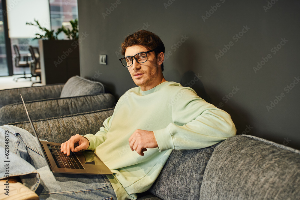 ambitious entrepreneur in stylish eyeglasses and casual clothes sitting on comfortable couch with laptop and looking at camera in contemporary office environment, confidence and success concept