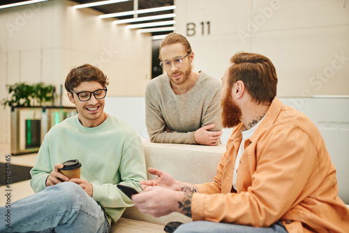 bearded man with mobile phone talking to smiling colleagues in eyeglasses while sitting on comfortable couch during coffee break in lounge of modern office, positive entrepreneurs discussing startup