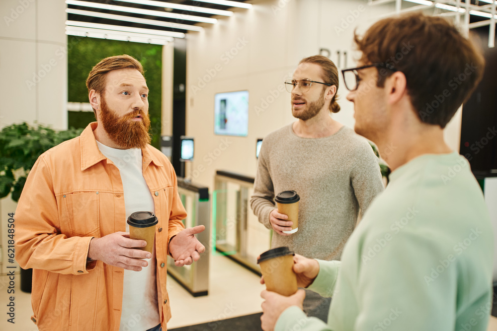 serious bearded man talking to colleagues in eyeglasses holding paper cups with takeaway drinks during coffee break in contemporary coworking office, partnership and collaboration in modern business serious bearded man talking to colleagues in eyeglasses holding paper cups with takeaway drinks during coffee break in contemporary coworking office, partnership and collaboration in modern business