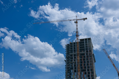 A construction crane and a building under construction against a blue sky with clouds. Construction site background. Cranes and new high-rise buildings