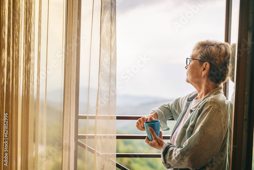 Beautiful senior elderly woman standing at the window overlooking the mountains and holding a mug of coffee in her hands