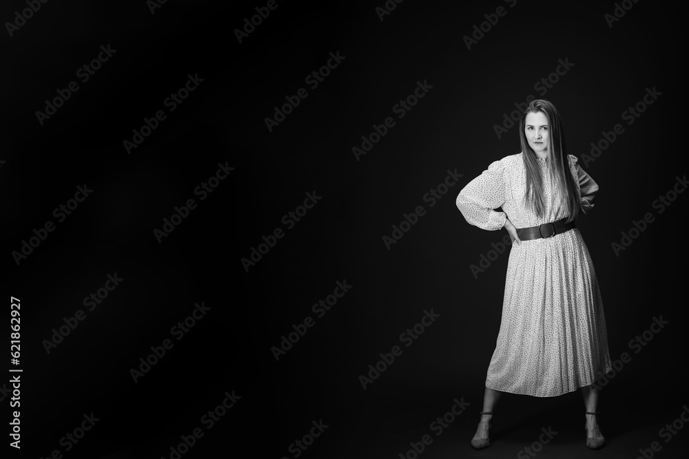 Young serious woman isolated on wall with copy space. Beautiful girl with hands on waist looking at camera. Portrait of lady in long dress looks confident on black background. Black and white photo.