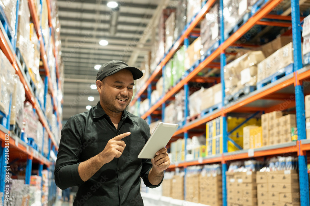 Warehouse worker wearing a hat and black shirt hands holding tablet check stock on tall shelves in warehouse storage. Asian auditor or staff work looking up stocktaking inventory in warehouse store. Warehouse worker wearing a hat and black shirt hands holding tablet check stock on tall shelves in warehouse storage. Asian auditor or staff work looking up stocktaking inventory in warehouse store.