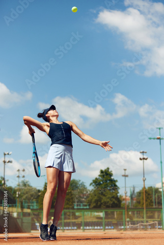 Full-length dynamic image of young athletic woman, tennis player in uniform during game, playing at open air stadium, court on warm day. Concept of sport, hobby, active lifestyle, health, strength, ad