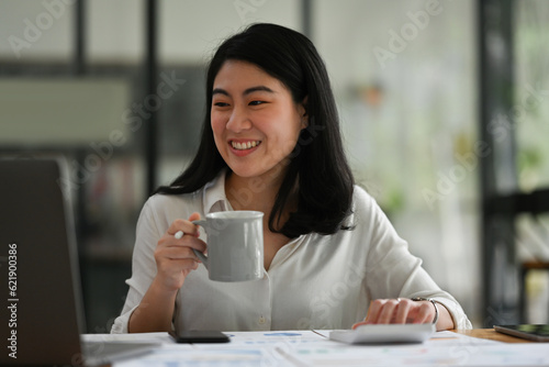 Photo of Young Asian accountant drinking coffee during numerical analysis on paperwork and laptop.