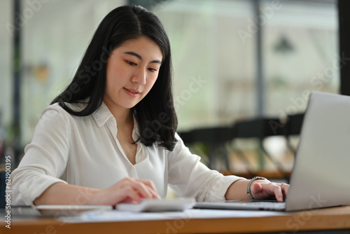 Portrait image of a young businesswoman or accountant pressing a calculator to make financial documents on laptop.