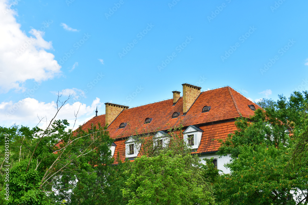 Rural house with white walls and red tiled roof in green trees on a sunny summer day with blue sky. Poland, Kurnik, Poznan, June 2022.
