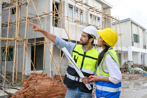 Young attractive construction man and woman in vests with helmets working on the under-construction building site. Home building project. Engineer foreman discusses with a coworker at workplace.