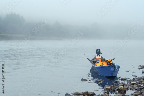 Fishing blue motorboat on a rocky river bank in the early morning in a foggy haze. High quality photo