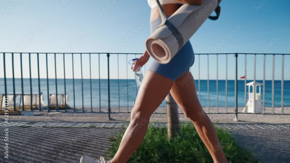 Athlete girl walking seacoast at twilight holding sport mat. Yoga woman going workout wearing sportswear outdoors. Attractive confident sportswoman enjoying training in beautiful summer evening