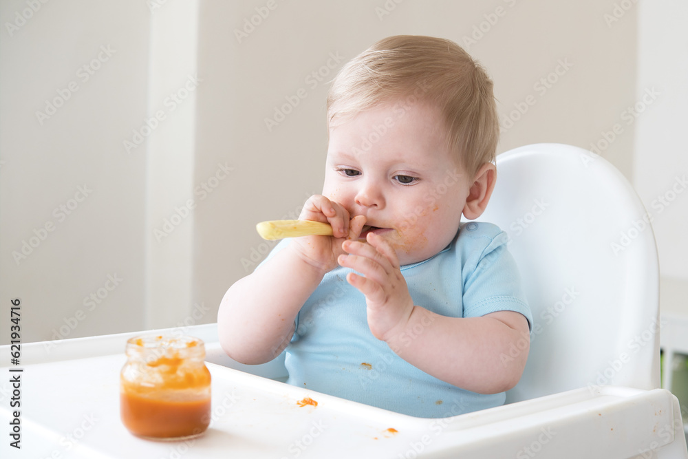 dirty baby boy holding spoon eating bland mashed food sitting, on high chair. first baby food dirty baby boy holding spoon eating bland mashed food sitting, on high chair. first baby food
