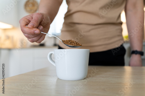 Woman pouring instant coffee granules into a white cup with a spoon. Girl with one spoonful of granulated coffee beans in a kitchen.