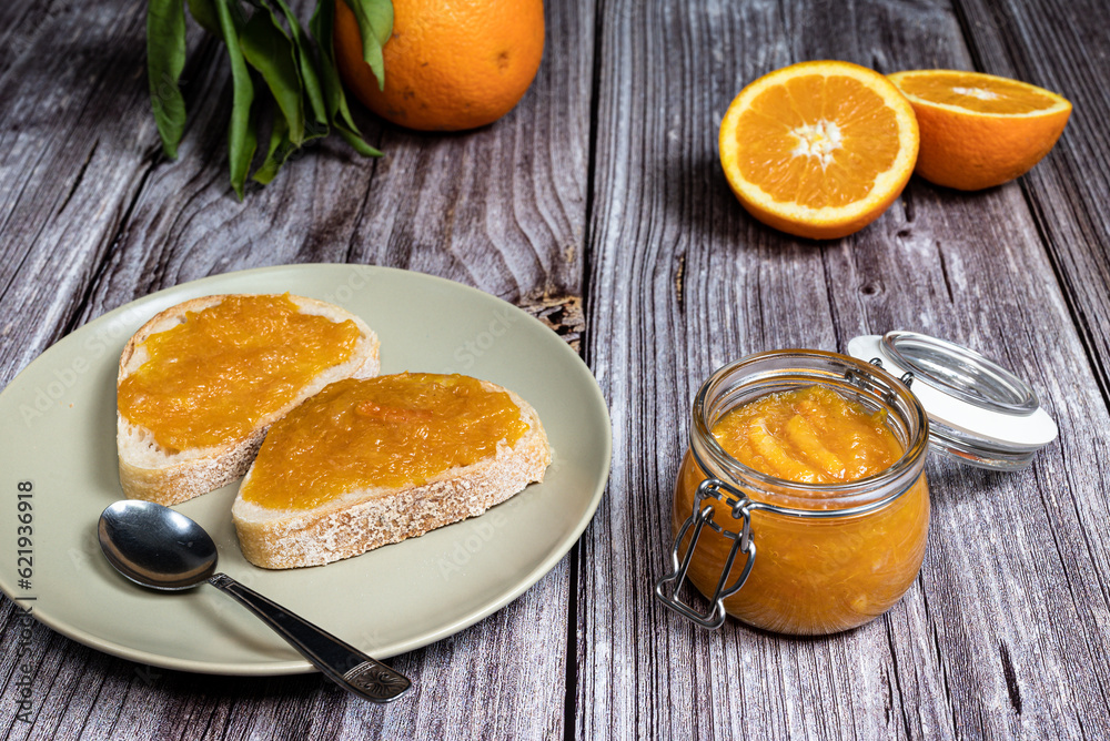 Delicious homemade orange marmalade in a glass jar. In the background slices of bread with marmalade in a plate on a white wooden table. Delicious homemade orange marmalade in a glass jar. In the background slices of bread with marmalade in a plate on a white wooden table.