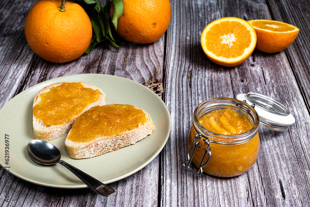 Delicious homemade orange marmalade in a glass jar. In the background slices of bread with marmalade in a plate on a white wooden table. Delicious homemade orange marmalade in a glass jar. In the background slices of bread with marmalade in a plate on a white wooden table.
