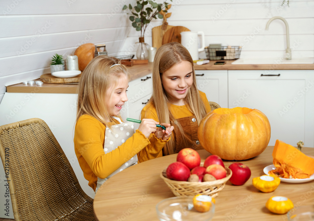 Two girls paint on a large orange pumpkin for a Halloween celebration while sitting at a wooden table at home. Family fun activity