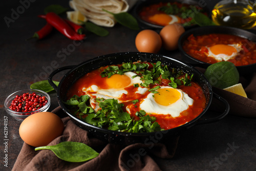 Pans with shakshuka, eggs, basil, bowl of pepper and oil on dark background