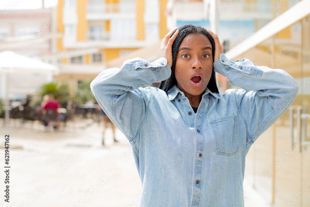 Fototapeta premium afro pretty black woman looking excited and surprised, open-mouthed with both hands on head, feeling like a lucky winner