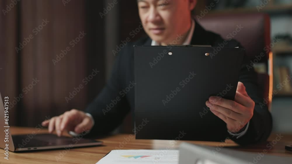 Close up of black clipboard and asian man looking at papers and then turning eyes on laptop screen. Successful businessman working at table in comfortable office space.