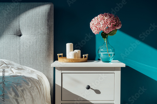 Stylish modern cosy bedroom in dark colors. Cozy interior with turquoise walls, home decor. Bed with grey fabric headboard, white blanket, bedside table, vase with pink hydrangea flower, candles.