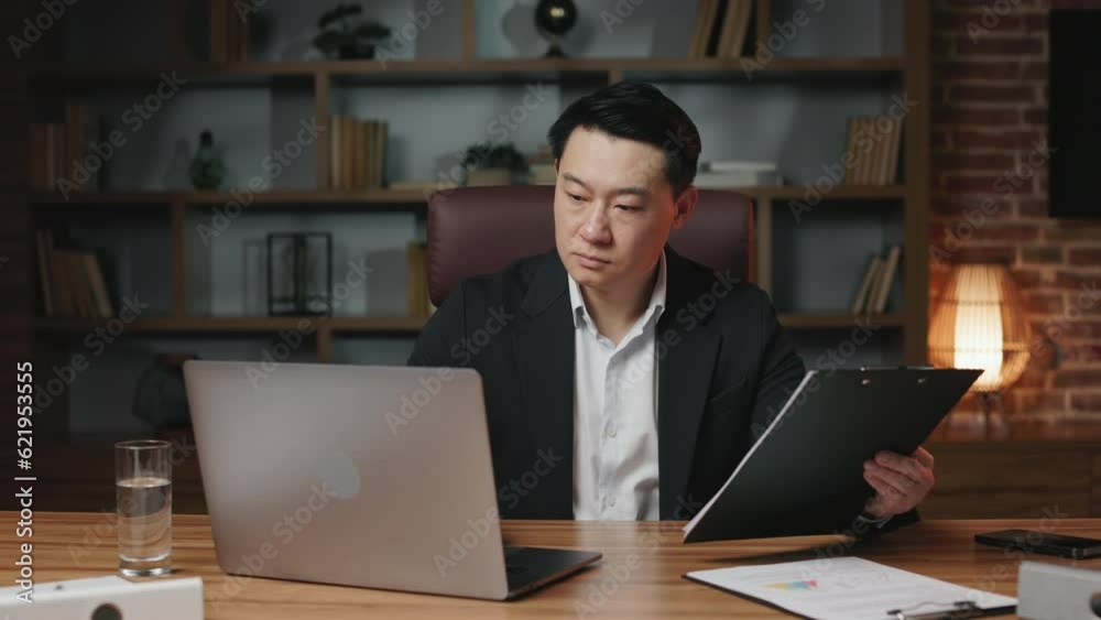 Diligent manager comparing paper contract with document received on corporate email. Asian man with serious face expression working in upscale office with remote computer. Diligent manager comparing paper contract with document received on corporate email. Asian man with serious face expression working in upscale office with remote computer.