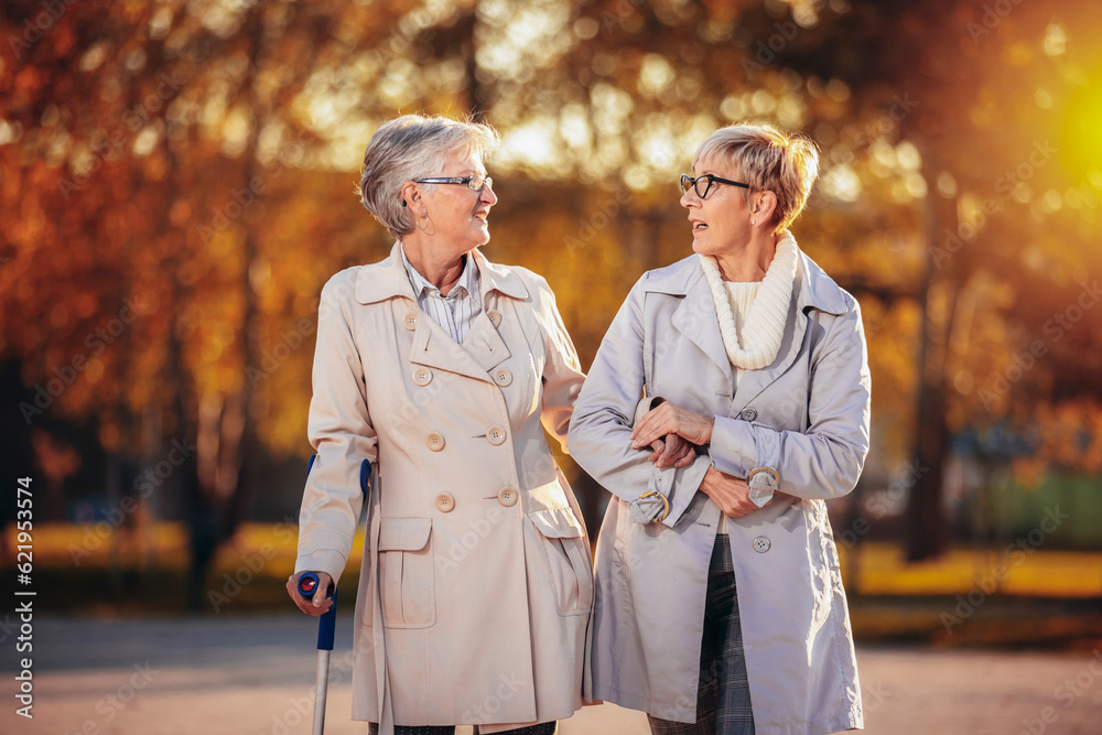 An older woman helping another woman that is walking with a cane in the park in the autumn An older woman helping another woman that is walking with a cane in the park in the autumn