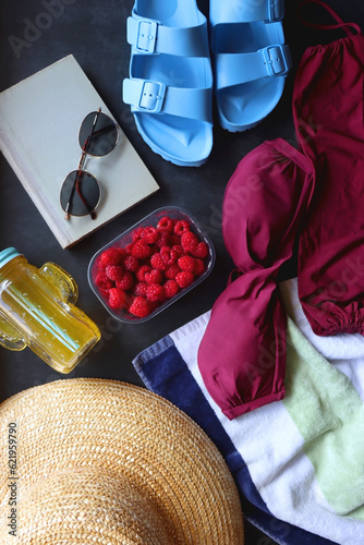 Maroon swimsuit, striped towel, straw hat, cactus cup with orange soda, bowl of raspberries, blue sandals, book and sunglasses. Summer or beach essentials. Top view, dark background.
