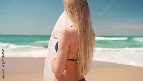 Surfing on travel vacation. Blonde woman in black bikini stands with surfboard in front beach and ocean looking camera. Healthy active lifestyle on summer vacation. Surfgirl on Atlantic Ocean photo