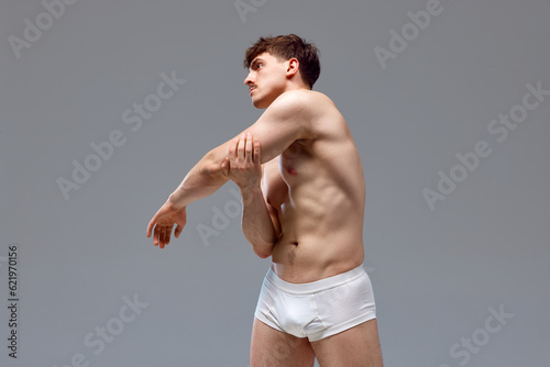 Back pains. Young man with relief, muscular, strong body, back posing shirtless in underwear against grey studio background. Concept of man's beauty, sport, health, athletic body, medicine photo