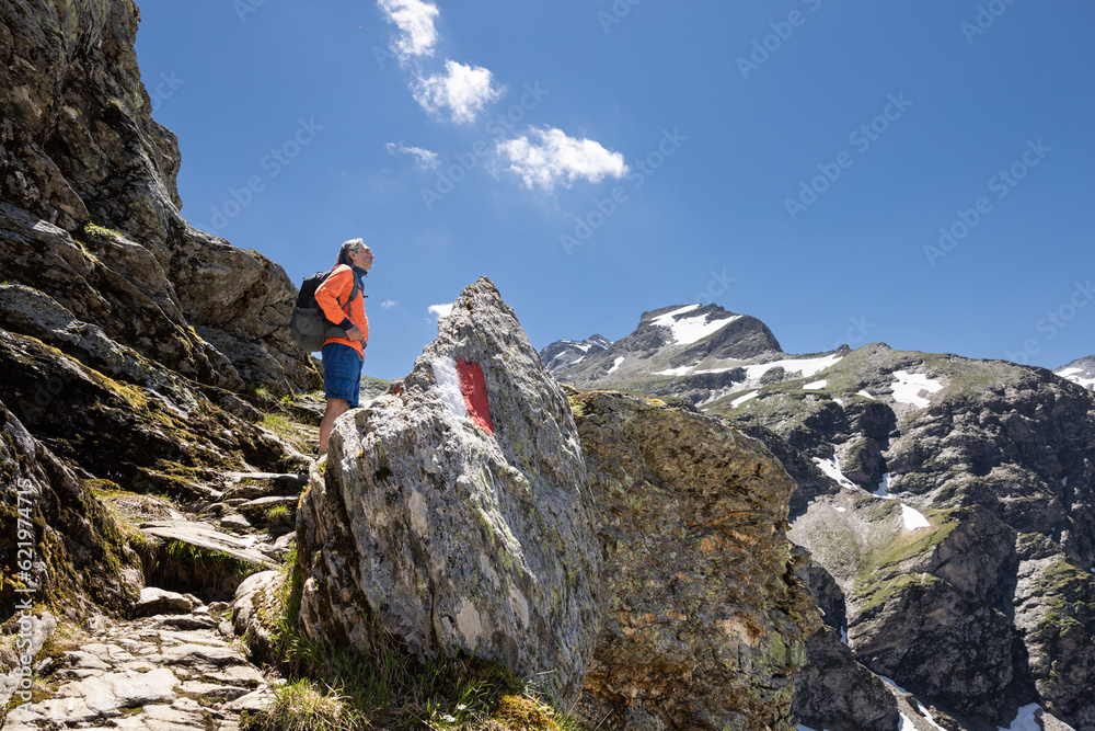 A gray-haired man with a backpack stands on a rocky mountain path with a route sign against the backdrop of rocks 