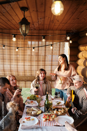 Thanksgiving Day, Family of five and dog Yorkshire terrier during family dinner outdoors at sunset table eating and drinking, having fun, singing songs with guitar