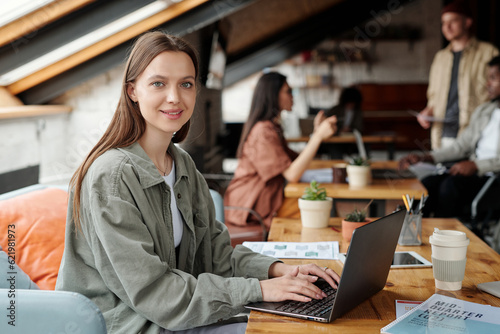 Young smiling female analyst or economist looking at camera while sitting by workplace in front of laptop and working with online financial data