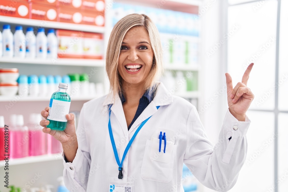 Blonde woman working at pharmacy drugstore holding syrup smiling happy pointing with hand and finger to the side
