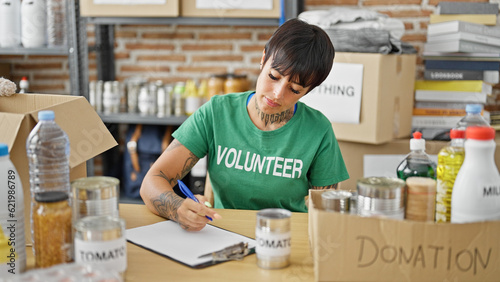 Young beautiful hispanic woman volunteer sitting on table writing on clipboard at charity center