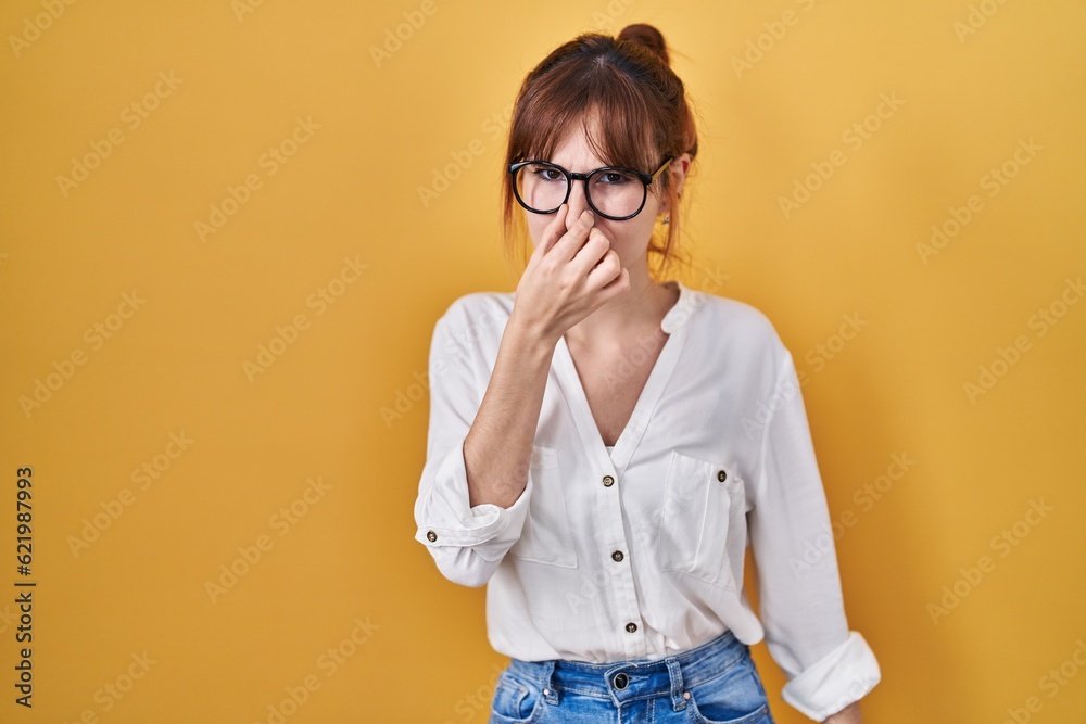 Young beautiful woman wearing casual shirt over yellow background smelling something stinky and disgusting, intolerable smell, holding breath with fingers on nose. bad smell Young beautiful woman wearing casual shirt over yellow background smelling something stinky and disgusting, intolerable smell, holding breath with fingers on nose. bad smell