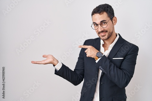 Handsome business hispanic man standing over white background amazed and smiling to the camera while presenting with hand and pointing with finger.