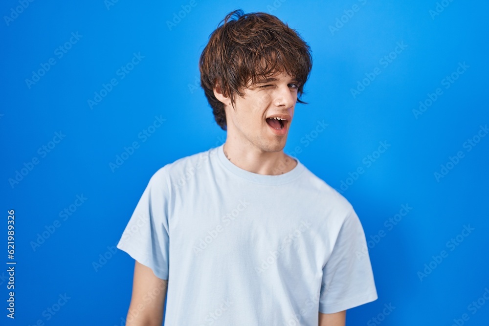 Hispanic young man standing over blue background winking looking at the camera with sexy expression, cheerful and happy face.