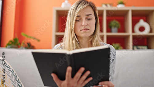 Young blonde woman reading book sitting on sofa at home