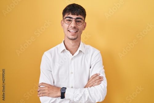 Young hispanic man standing over yellow background happy face smiling with crossed arms looking at the camera. positive person.