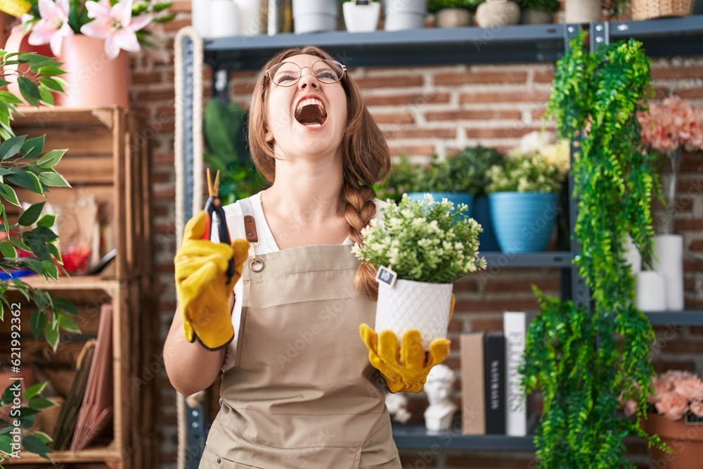 Young caucasian woman working at florist shop holding plant angry and mad screaming frustrated and furious, shouting with anger looking up. Young caucasian woman working at florist shop holding plant angry and mad screaming frustrated and furious, shouting with anger looking up.