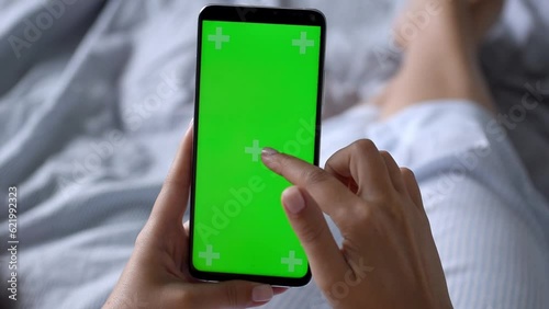 The girl holds a smartphone with a green screen in her hands. Over the shoulder view from the back of a girl lying on the bed and flipping through the smartphone screen. Relaxed home mood
